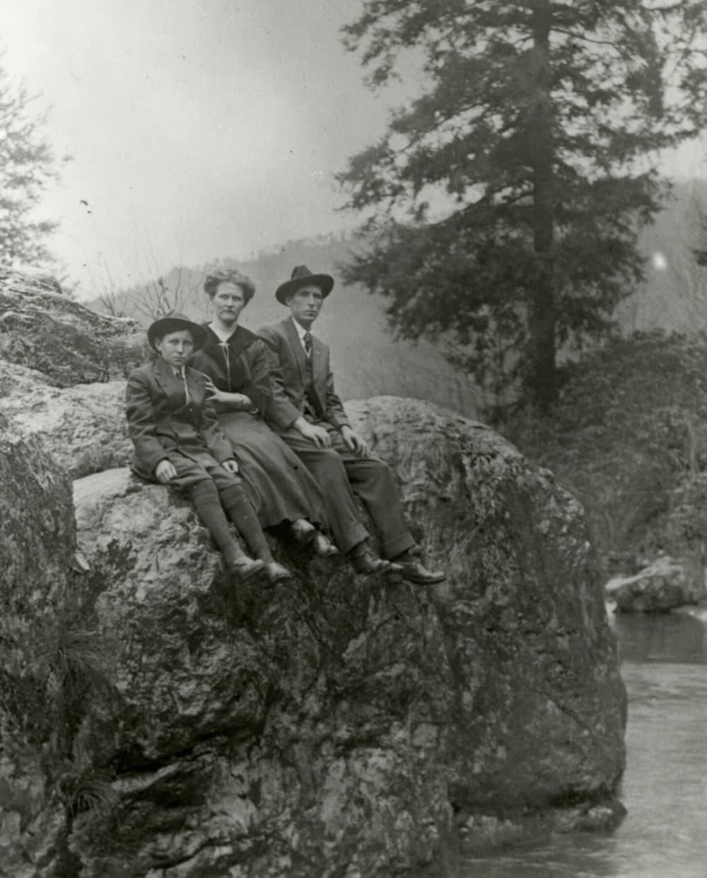 man woman and boy sitting on large rock by creek